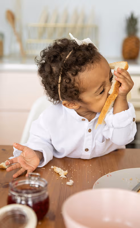 Baby girl eating her heart out of a toast, crumbs all over the table. Shot by KATRIN BOLOVTSOVA