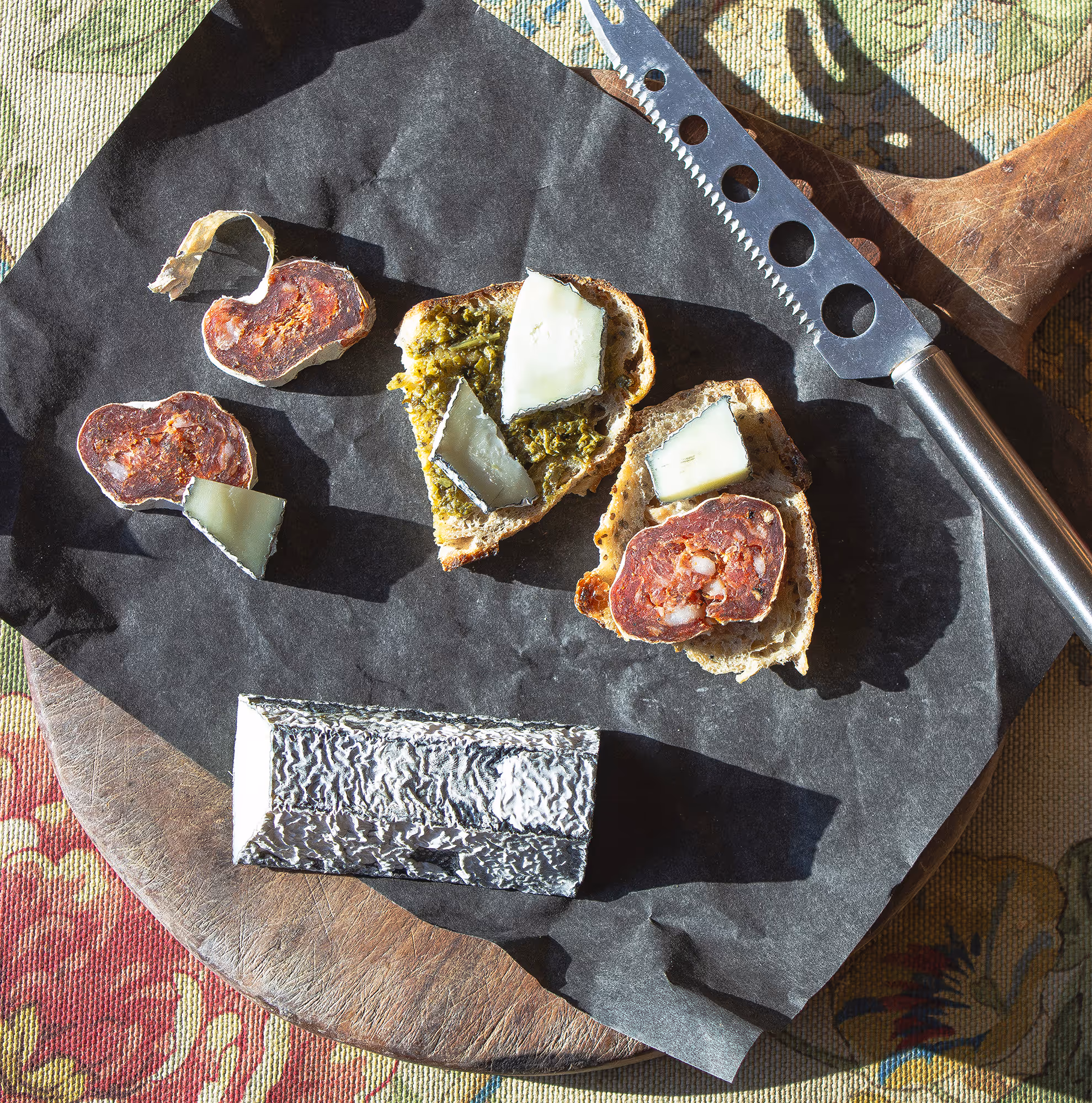 Bird's eye view of a charcuterie wooden board in the afternoon sun on a table cloth, Black Charcuterie paper, cheese knife, cut saucisson, pesto'd toasts with goat cheese on top. 
