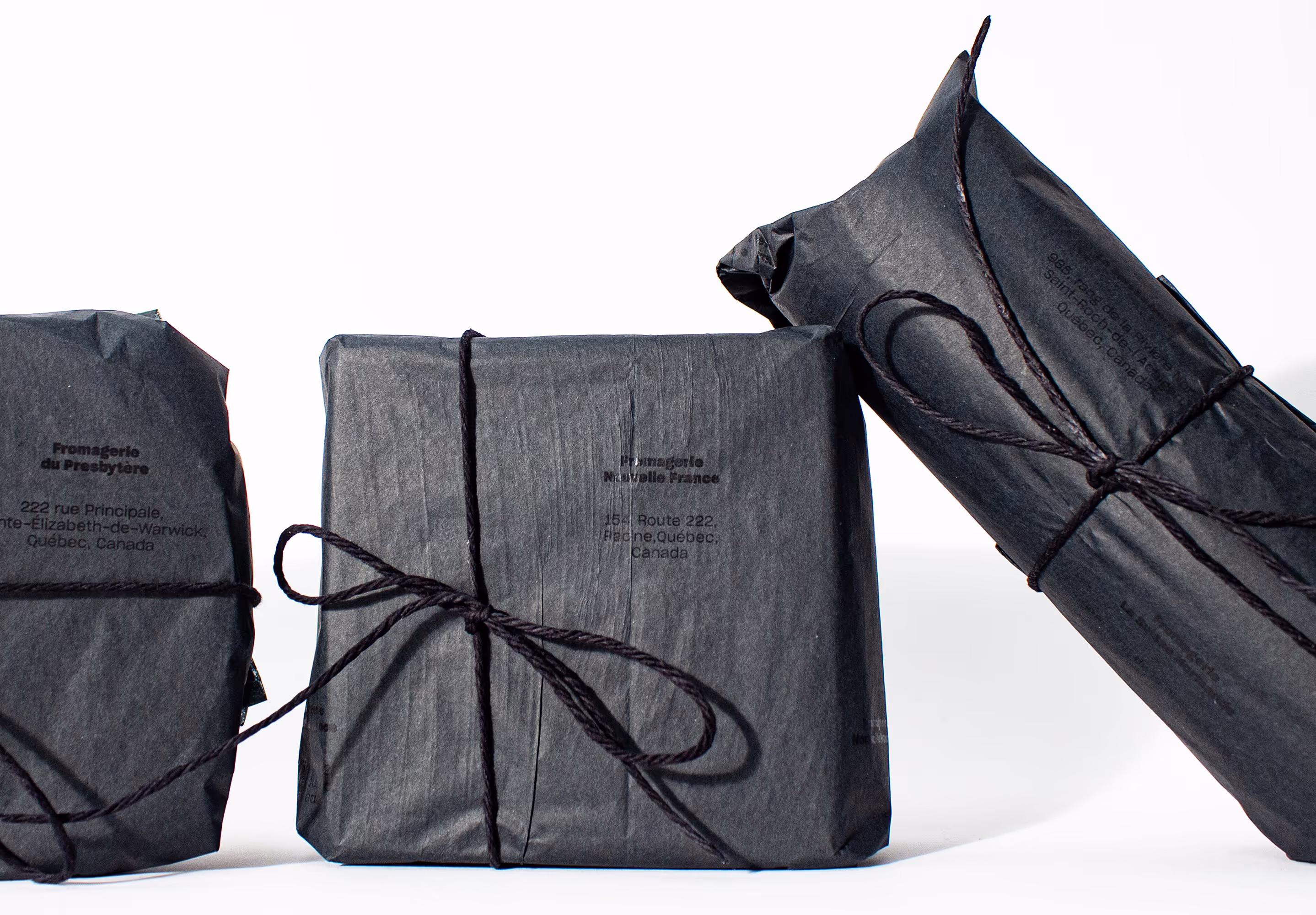 Horizontal studio shot of the three cheeses with only their first layer of black charcuterie paper, wrapped by black strings. Locations of the original farms printed black on black.