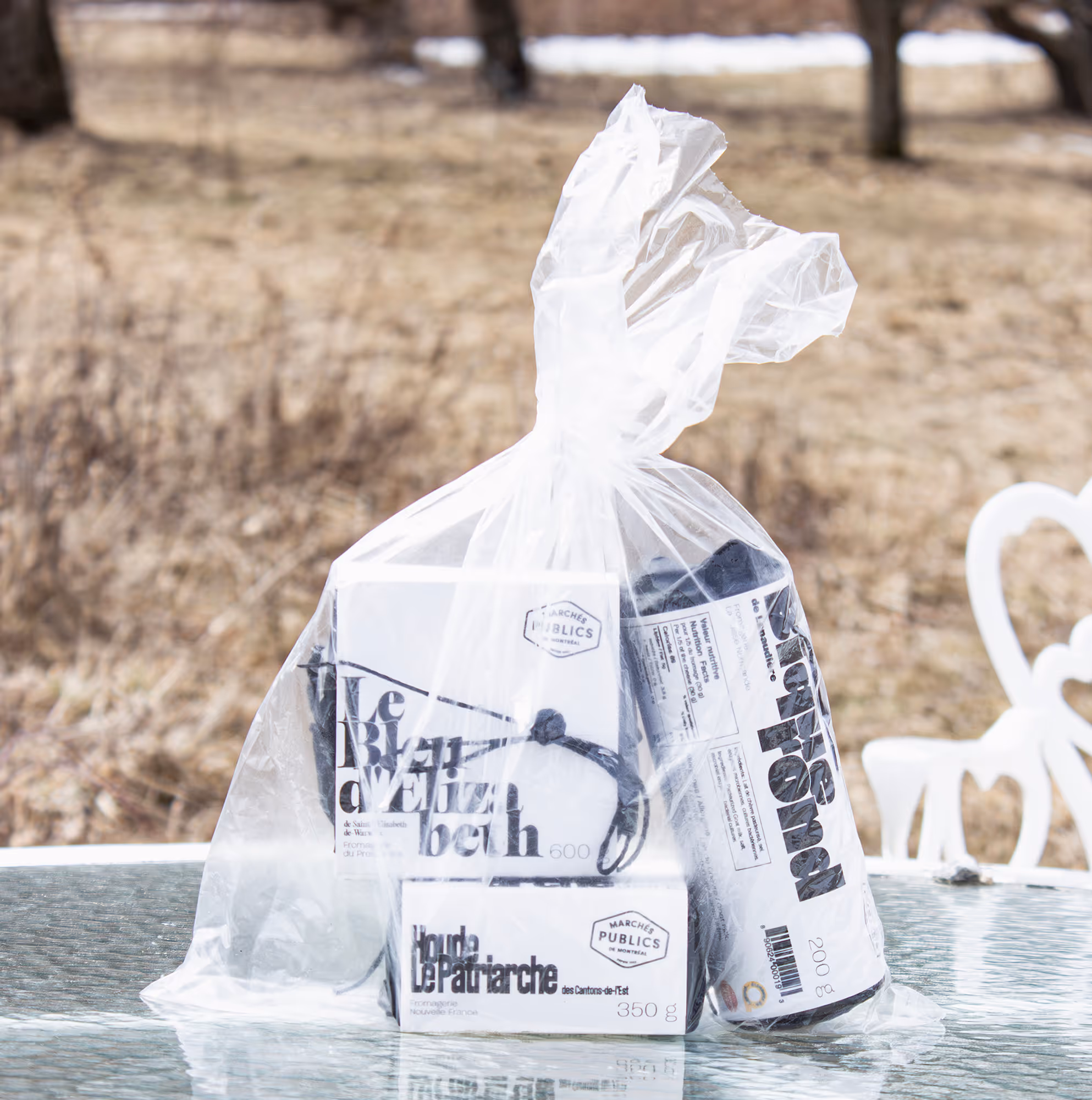 Detailed zoom in on a plastic bag holding all three packaged cheeses. Bag sitting on a white table outside, rural and cloudy muted background.