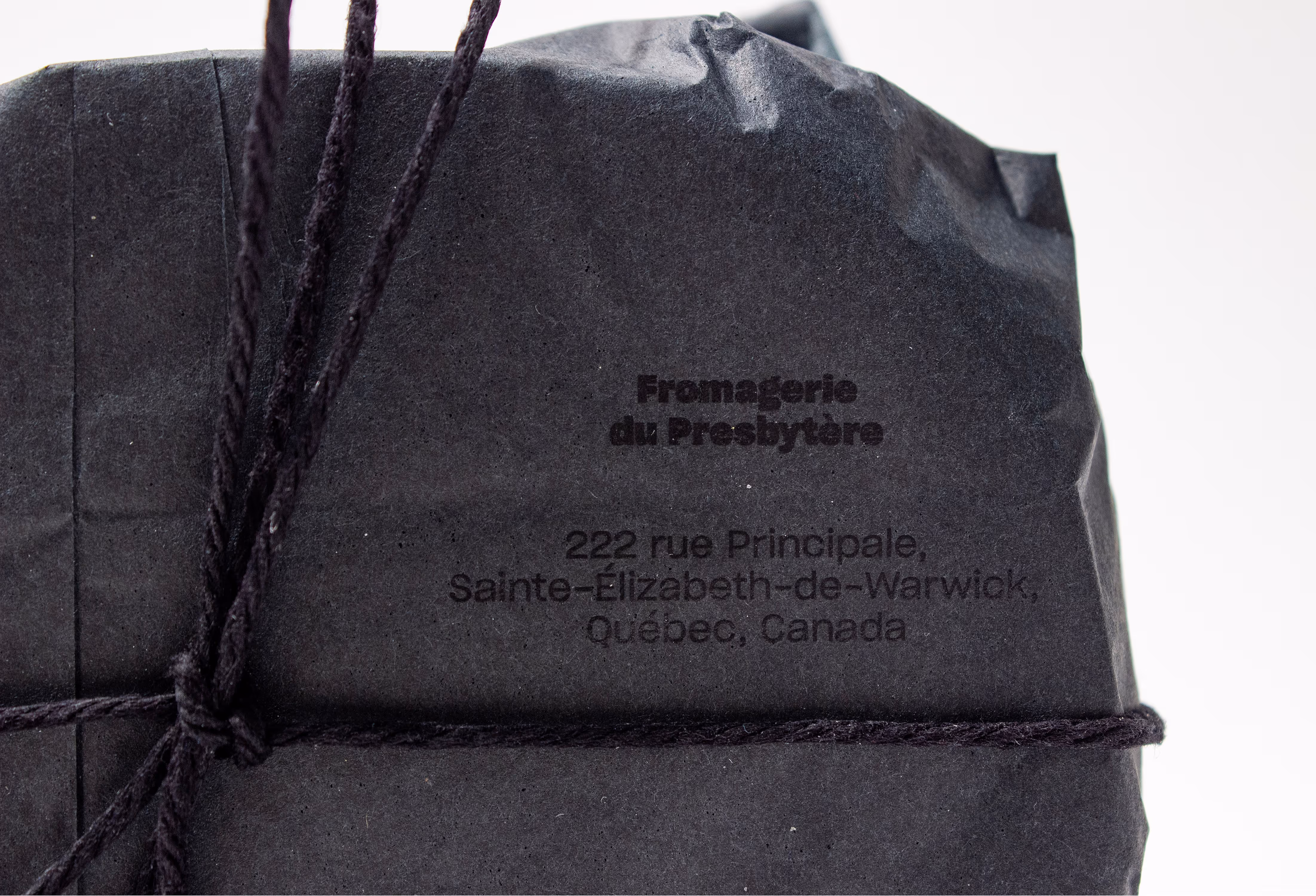 high quality close-up of the un-sleeved black packaging. "Fromagerie du Presbitère — 222 rue Principale, Sainte Élizabeth de Warwick, Québec, Canada" printed on black on the lighter black paper wrap.