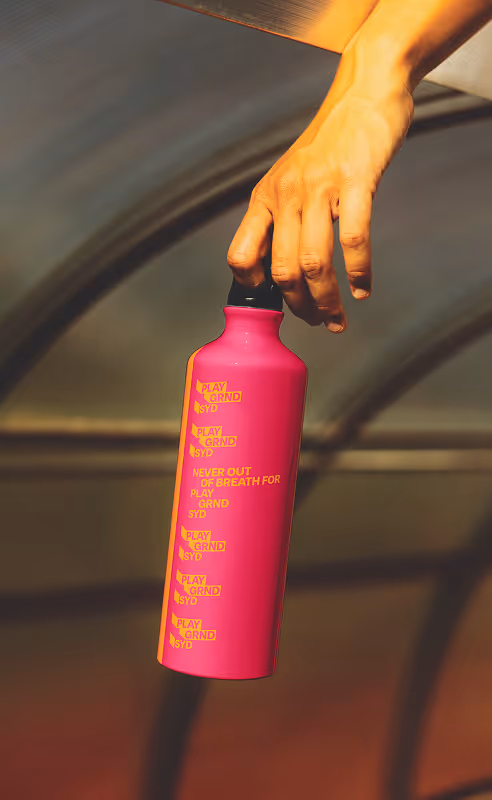 Hand holding a pink water bottle with orange tagline and logos "never out of breath for play grnd syd". Source : Mockup Maison