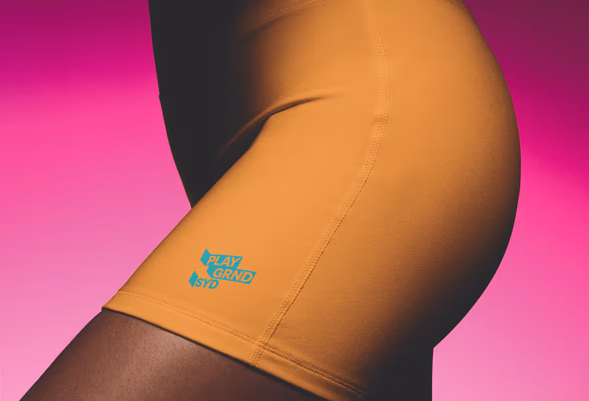 Close up of woman wearing Orange yoga shorts with blue Sydney (SYD) logo near the hem. Pink Background. Source : Mockup Maison